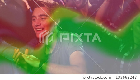 Smiling man in green shirt clapping on club dance floor, with wristwatch and colored light streaks 135668248