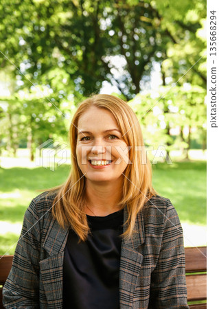 A portrait of a smiling middle-aged woman with blonde hair sitting on a wooden bench in a park. Vertical portrait woman 40s years A portrait of a smiling middle-aged woman with blonde hair sitting on a wooden bench in a park. Vertical portrait woman 40s years 135668294