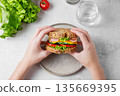 A hands holding a sandwich with ham, cheese, lettuce, and vegetable over a plate  135669395