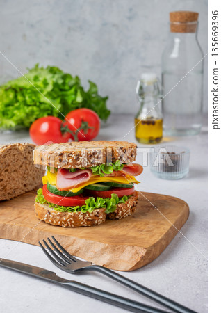 Sandwich with ham, cheese, lettuce and vegetable on a wooden board on a light background Sandwich with ham, cheese, lettuce and vegetable on a wooden board on a light background 135669396