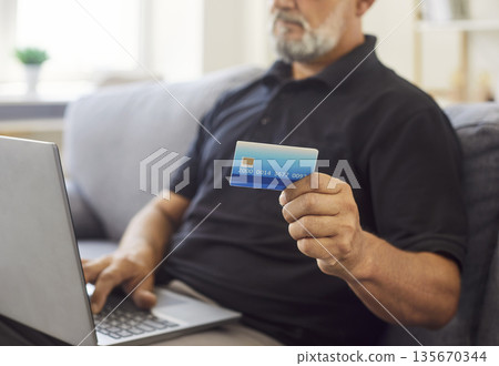Senior man holding credit card while using laptop for online payment at home Senior man holding credit card while using laptop for online payment at home 135670344