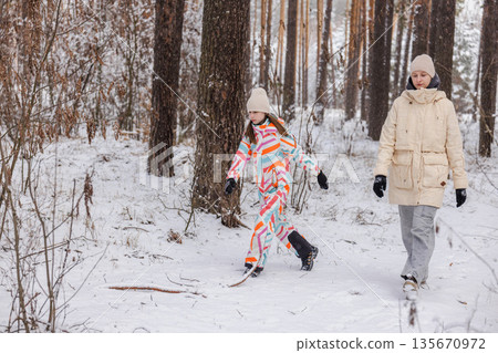Two teenagers walking along snowy forest path in winter, outdoor leisure scene with people in pine woodland, cold weather atmosphere 135670972