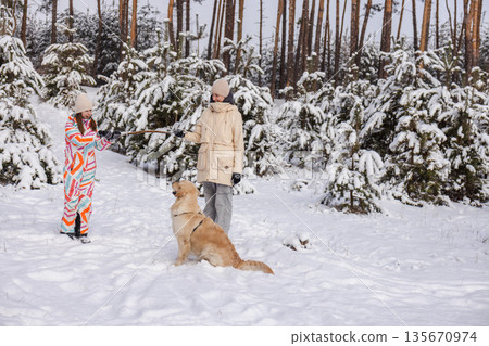 Two teenagers playing tug with Golden Retriever in snowy pine forest, winter outdoor leisure, joyful family moment 135670974