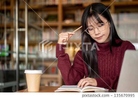 Glasses asian woman holding pencil and thinking beside laptop and coffee on wooden table in library. 135670995