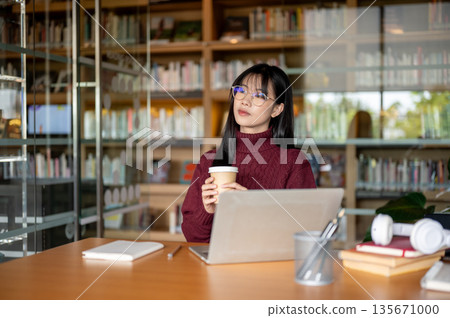 Glasses asian woman holding coffee looking away while sitting with laptop at wooden table in library 135671000
