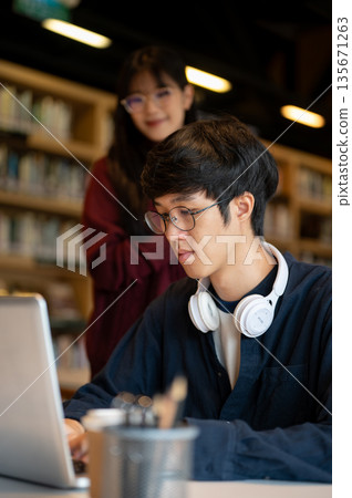 Glasses asian man student looking at laptop with a woman friend standing beside table in library. Glasses asian man student looking at laptop with a woman friend standing beside table in library. 135671263