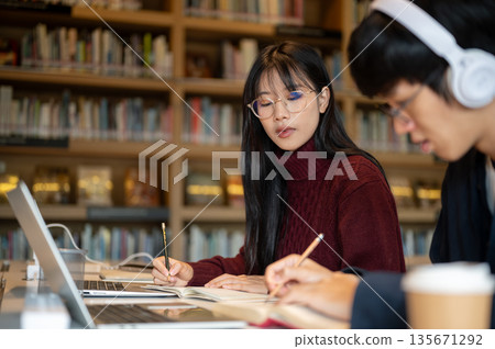 Glasses asian woman student looking at man friend writing in notebook while sits at library's table. Glasses asian woman student looking at man friend writing in notebook while sits at library's table. 135671292