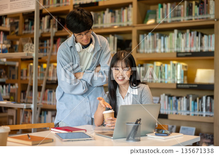 Glasses asian man student cross arms standing behind woman friend looking at laptop on library table 135671338