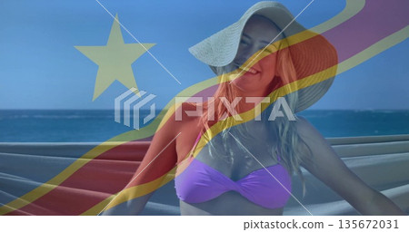 Standing woman wearing straw sun hat and purple bikini top on seaside terrace, with flag overlay Standing woman wearing straw sun hat and purple bikini top on seaside terrace, with flag overlay 135672031