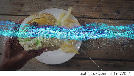 Lifting sesame-seed bun burger with adult hand over wooden tabletop, with ceramic plate of fries Lifting sesame-seed bun burger with adult hand over wooden tabletop, with ceramic plate of fries 135672065