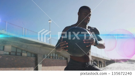 Shirtless man performing twisting exercise with black ball and smartwatch on beach walkway 135672240