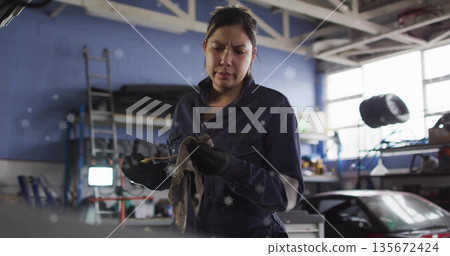 Hispanic mechanic wiping engine oil dipstick with rag in car repair garage, with ladder spare tire 135672424