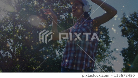 Woman wearing red blue shirt, baseball cap and over-ear headphones dancing in park, with sparkles Woman wearing red blue shirt, baseball cap and over-ear headphones dancing in park, with sparkles 135672480