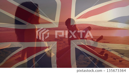 Sitting couple wearing swimwear on sandy beach at sunset, with surfboards and Union Jack overlay 135673364