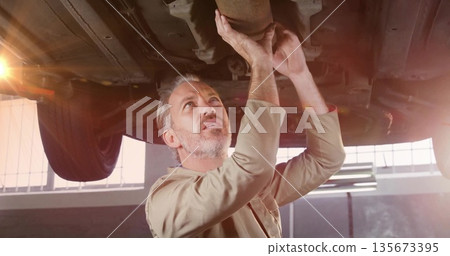 Inspecting catalytic converter, mechanic in coveralls reaching beneath vehicle on lift in garage Inspecting catalytic converter, mechanic in coveralls reaching beneath vehicle on lift in garage 135673395