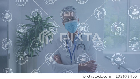 Standing doctor crossing arms wearing surgical mask and stethoscope in clinic, with digital icons 135673410