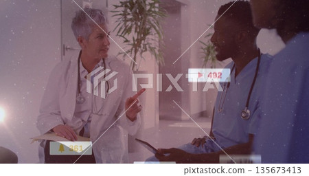 Gesturing senior doctor speaking with colleague in hospital corridor, with tablet and alert icons 135673413