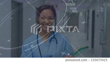Smiling nurse wearing scrubs and stethoscope standing in hospital corridor, holding patient folders 135673425