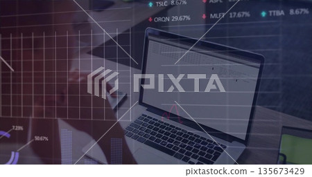 Mid-adult woman typing on silver laptop holding smartphone at desk, with floating chart overlays 135673429