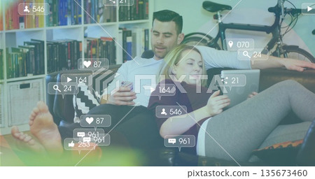 Reclining couple holding smartphone and tablet on living room sofa, with bookshelf and bicycle 135673460