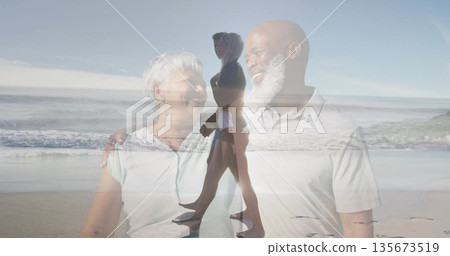 Mature couple laughing and looking at each other on beach, overlaying walking woman holding device 135673519