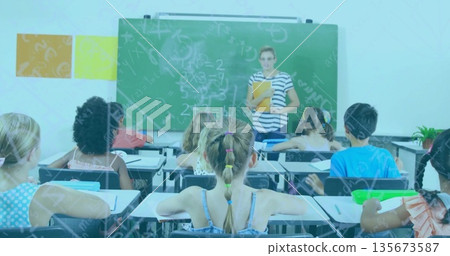 Teacher explaining math on chalkboard to children, wearing striped top and holding folder in school 135673587