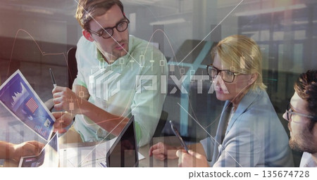 Finance team analyzing market data around conference table at office, with charts and laptops 135674728