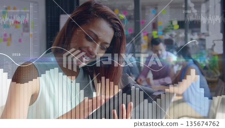 Smiling auburn-haired teammate tapping tablet in office, with laptops sticky notes data overlays Smiling auburn-haired teammate tapping tablet in office, with laptops sticky notes data overlays 135674762