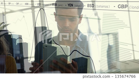Analyst in denim shirt analyzing market data on tablet in office, with charts and currency tickers 135674766