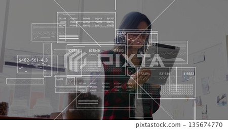 Analyzing woman interacting with floating data interfaces in office, with tablet and laptop 135674770