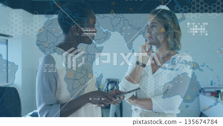 Discussing data businesswomen wearing suits using tablet at office desks, with map overlay monitors Discussing data businesswomen wearing suits using tablet at office desks, with map overlay monitors 135674784