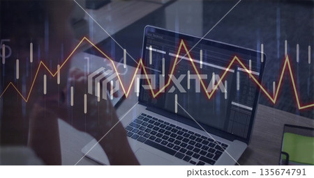 Reviewing chart data on laptop, woman wearing light shirt at wooden desk in office, with smartphone 135674791