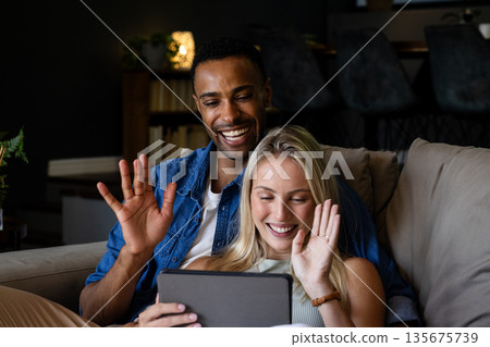 Couple on couch video chatting on tablet, smiling and waving at home 135675739