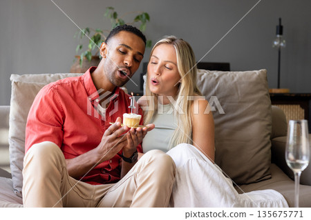 Couple celebrating birthday at home, blowing candle on cupcake together Couple celebrating birthday at home, blowing candle on cupcake together 135675771