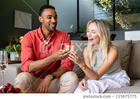 Man giving gift to smiling woman on cozy sofa at home, celebrating together Man giving gift to smiling woman on cozy sofa at home, celebrating together 135675775