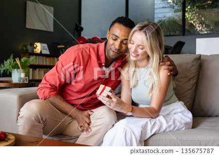 Smiling couple exchanging gift on sofa, enjoying joyful moment together Smiling couple exchanging gift on sofa, enjoying joyful moment together 135675787