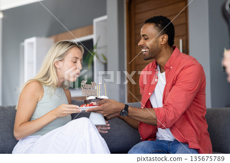 Celebrating birthday, woman blowing candles on cake while man smiling at home Celebrating birthday, woman blowing candles on cake while man smiling at home 135675789