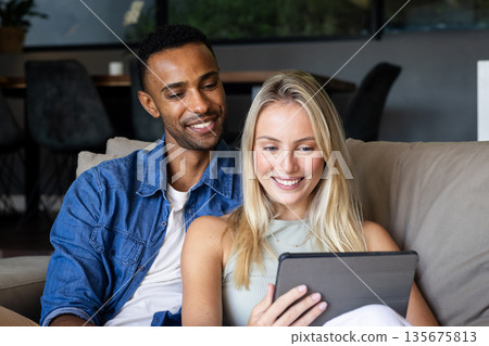Couple relaxing on couch, smiling while watching something on tablet at home Couple relaxing on couch, smiling while watching something on tablet at home 135675813