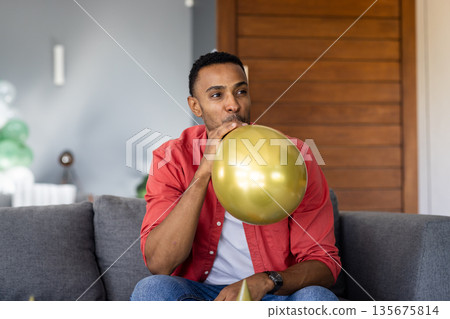 Man inflating gold balloon on couch, preparing for festive celebration Man inflating gold balloon on couch, preparing for festive celebration 135675814
