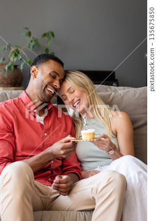 Couple on sofa sharing cupcake, laughing and enjoying cozy celebration at home 135675850