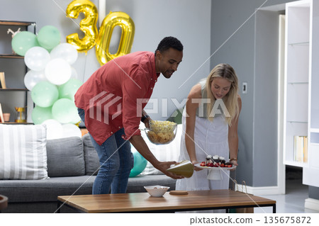 Celebrating birthday, couple arranging cake and snacks in living room with balloons 135675872