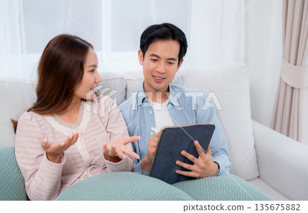 Happy asian couple planning and writing notes on digital tablet device together sitting on sofa. 135675882