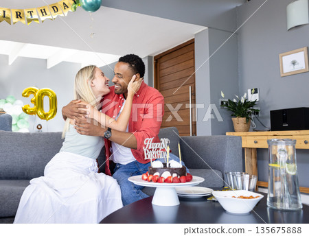 Couple celebrating birthday at home, embracing with cake and decorations around Couple celebrating birthday at home, embracing with cake and decorations around 135675888