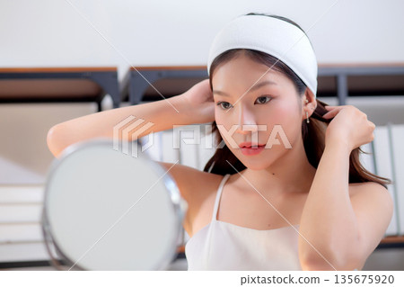 Young asian woman wearing headband and tying hair while preparing skincare looking mirror. 135675920