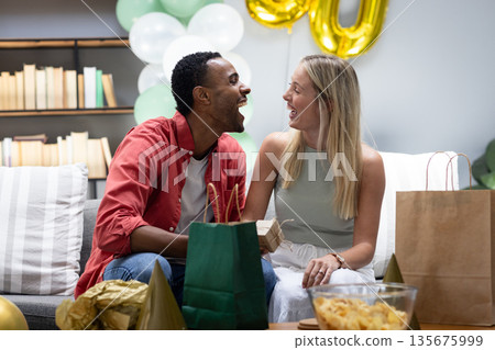 Couple joyfully exchanging gifts at home during festive celebration with balloons 135675999