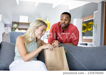 Couple celebrating at home, woman opening gift bag with excitement and joy 135676000