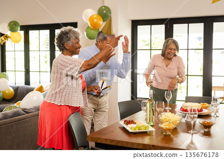 Elderly friends celebrating at home, enjoying food and drinks with laughter 135676178