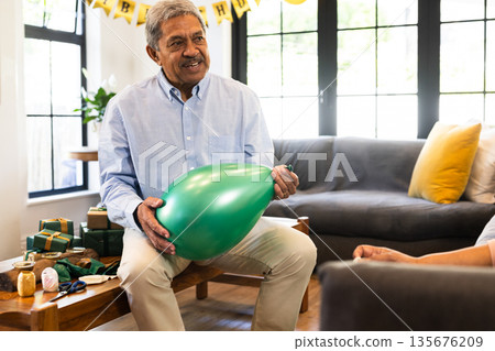 Elderly man inflating balloon at home, preparing for celebration with gifts Elderly man inflating balloon at home, preparing for celebration with gifts 135676209