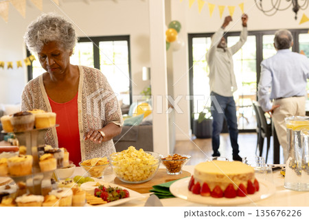 Elderly woman arranging cupcakes on table while guests celebrating in background Elderly woman arranging cupcakes on table while guests celebrating in background 135676226