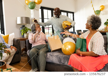 Elderly friends celebrating birthday at home with gifts and balloons, sharing joy Elderly friends celebrating birthday at home with gifts and balloons, sharing joy 135676237